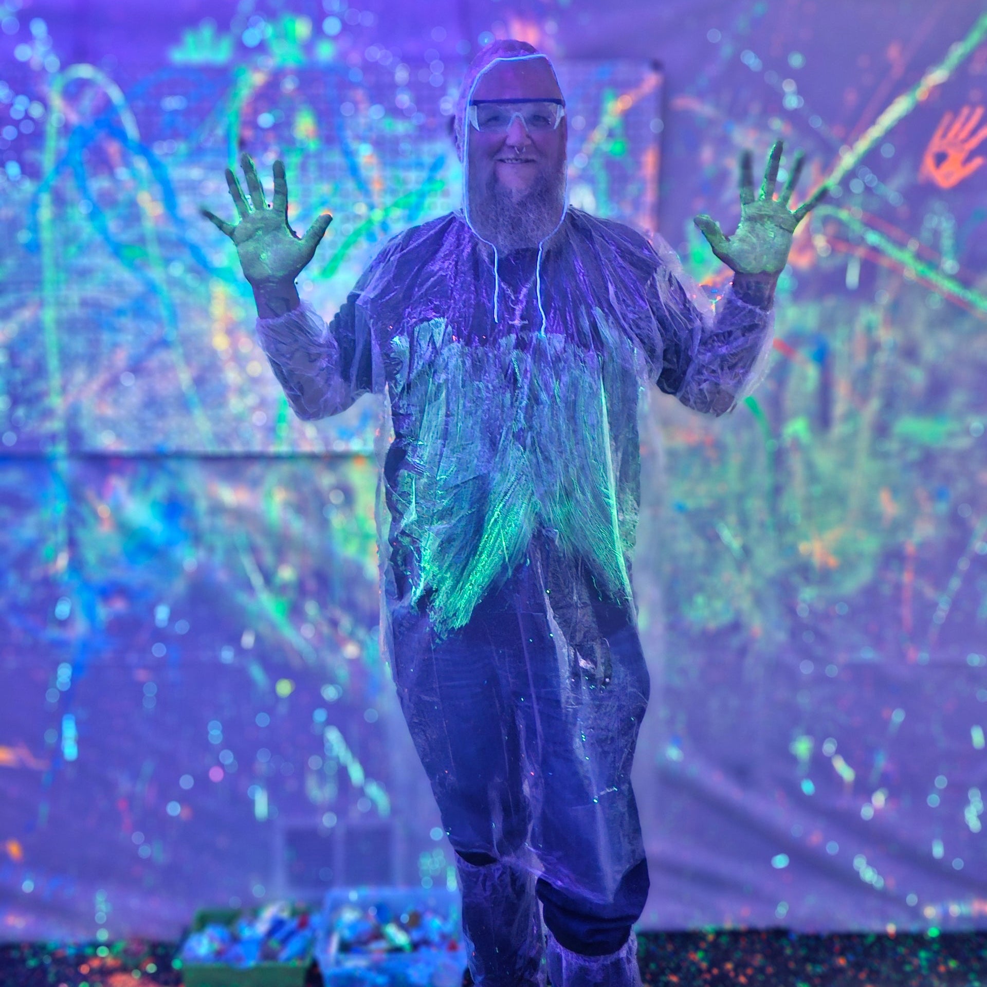 A man in a protective poncho, glasses, and booties stands in as paint splattered room and shows off his painted hands after playing in the Splatter Spot UV-reactive splatter painting room.